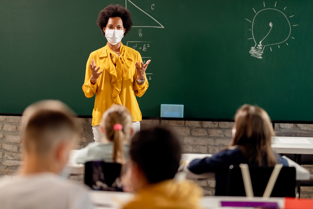Teacher standing by chalkboard wearing a mask at the head of the classroom